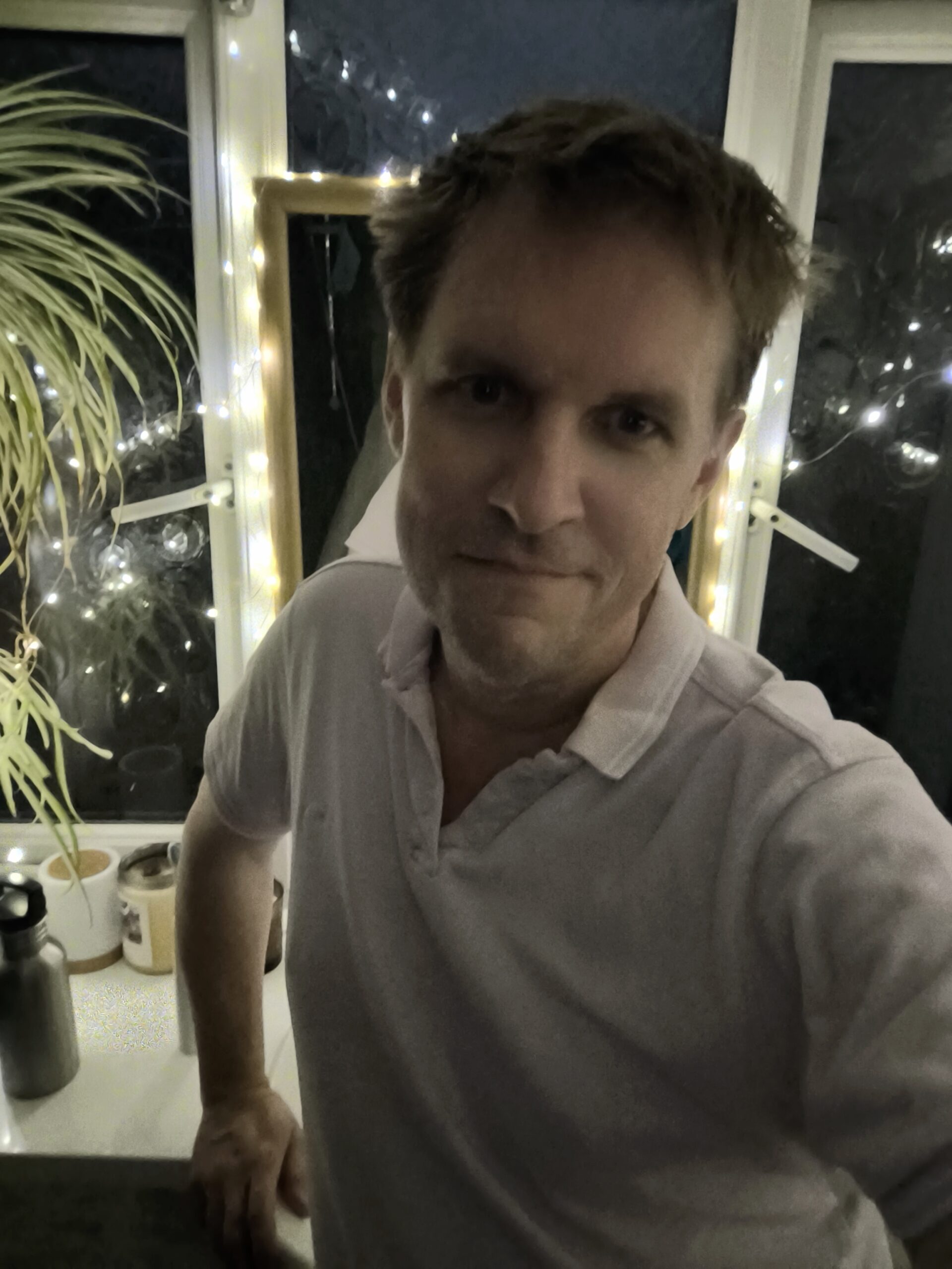 Patrick leaning casually against a windowsill, smiling slightly, with fairy lights glowing in the background. Personal photo for About page