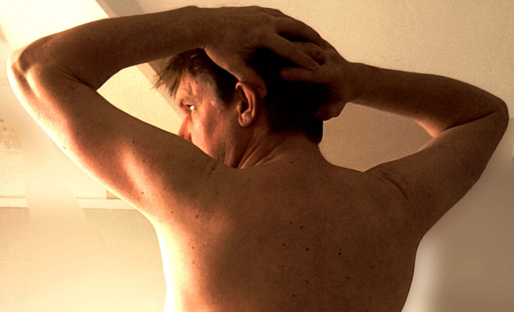 Black and white photo of Patrick with arms raised behind his head, bare-backed, representing vulnerability and submission.